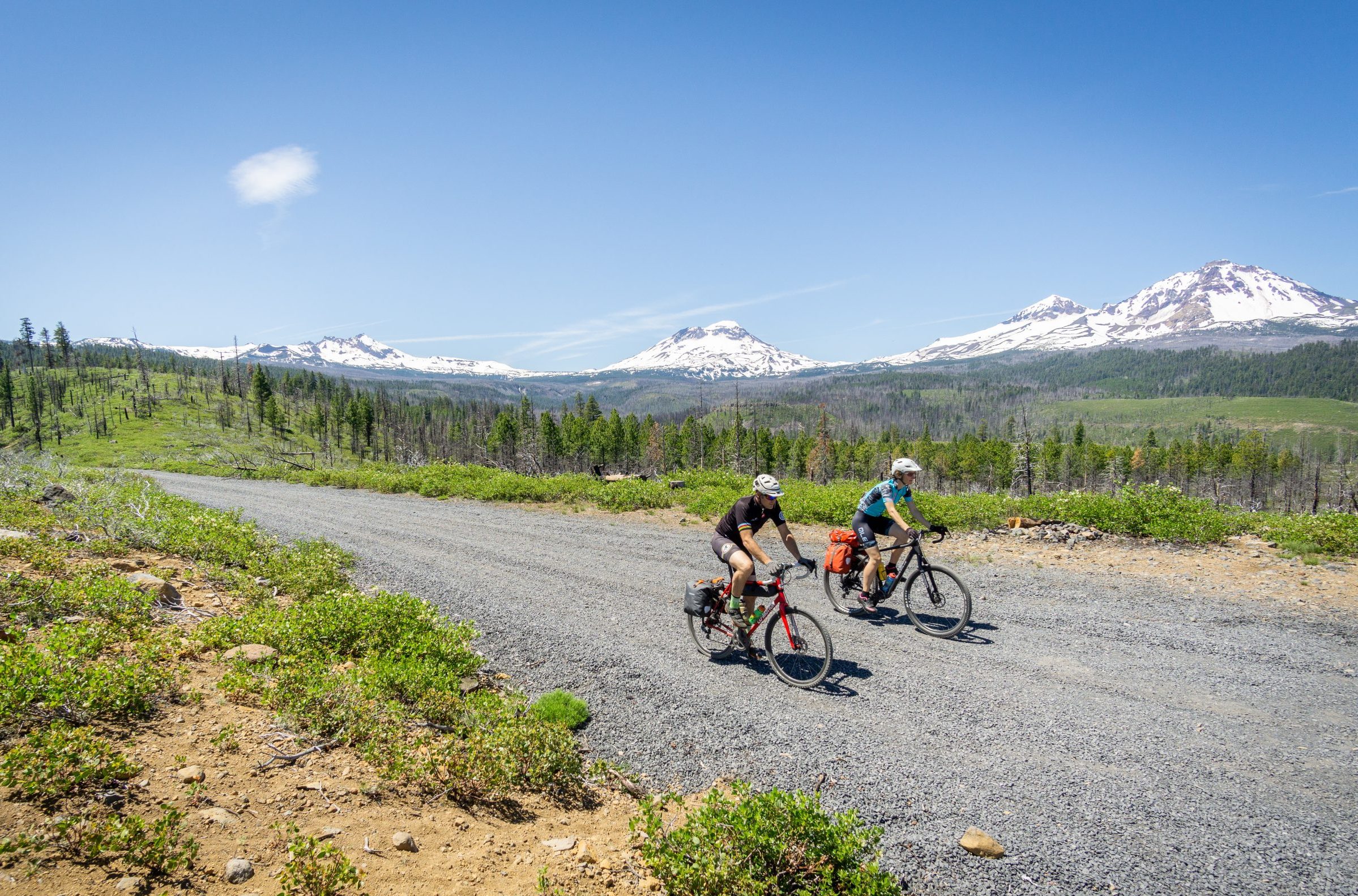 OMM_GRAVEL_6000p-6 Two cyclists riding on a gravel road with the Sisters mountains in the background.